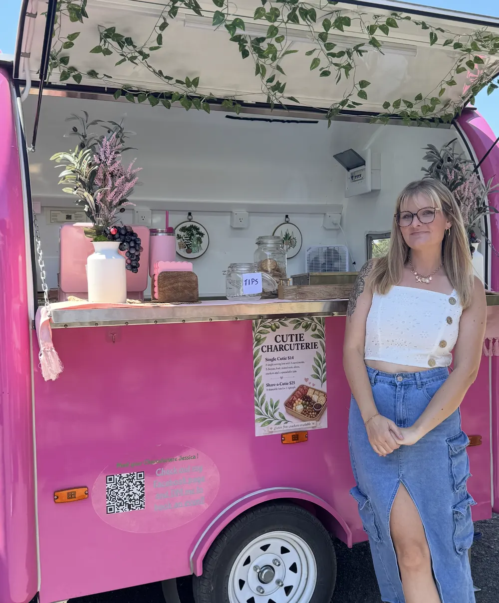 The pink Cutie Charcuterie trailer with menu sign and Jessica out front
